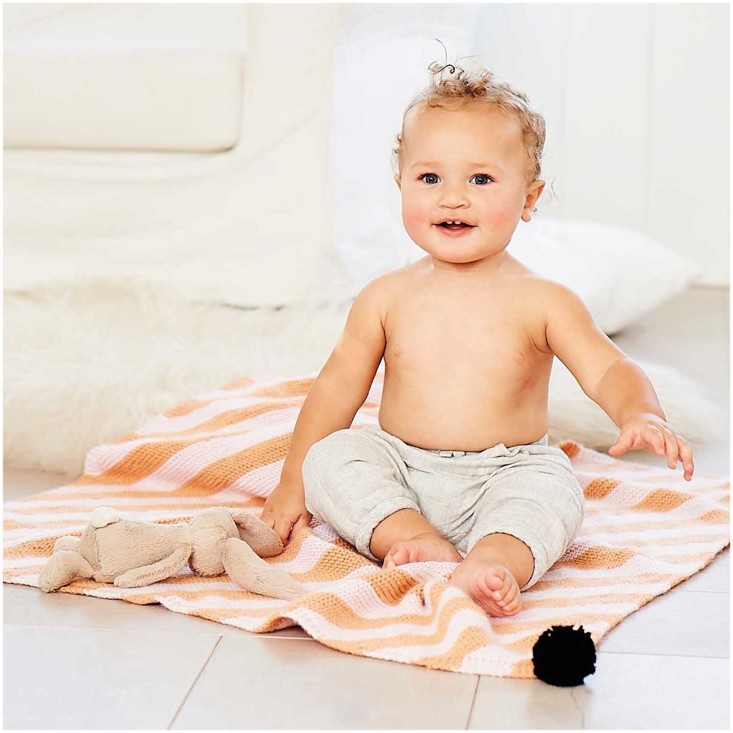 Rico Baby Dream dk uni - A Luxury Touch: Ein lächelndes Baby sitzt auf einer rosa Decke von Rico Design.
