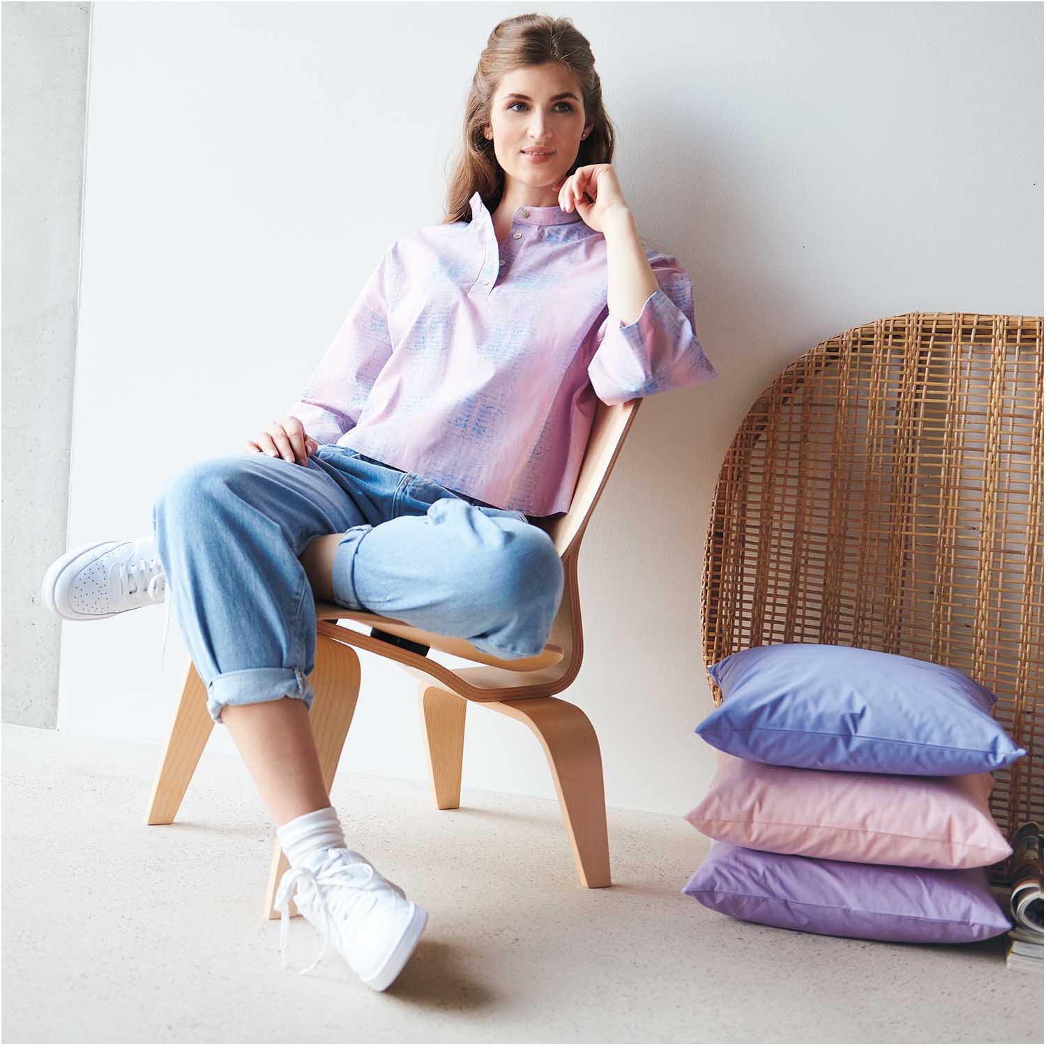 Stoffabschnitt Baumwollstoff uni lila 50x140cm: Frau in lässiger Kleidung sitzt auf Stuhl, daneben lila und rosa Kissen, modernes Design.