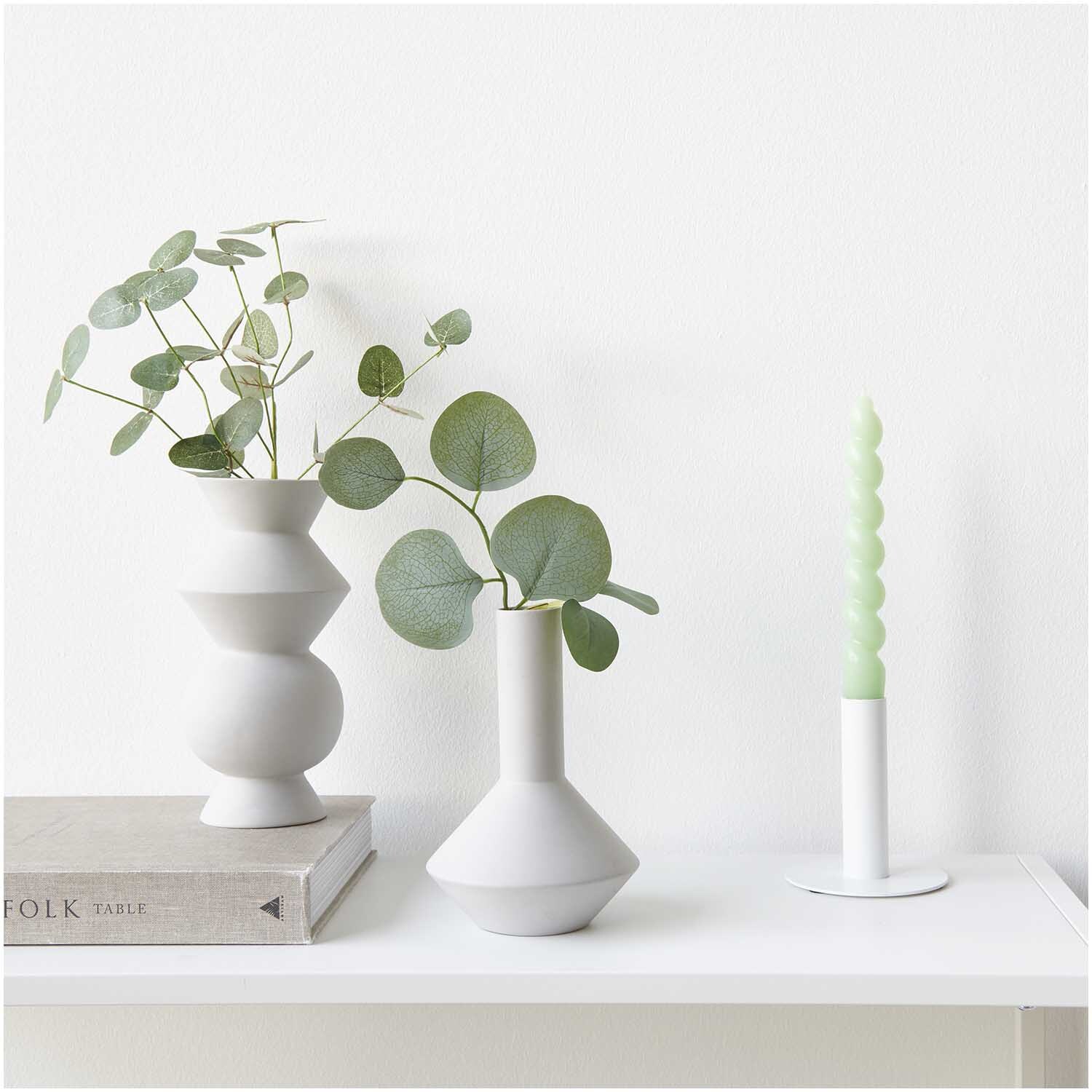 Eukalyptuszweig 20cm in eleganter Vase neben einer grünen Kerze auf einem weißen Regal. Marke: Rico Design.