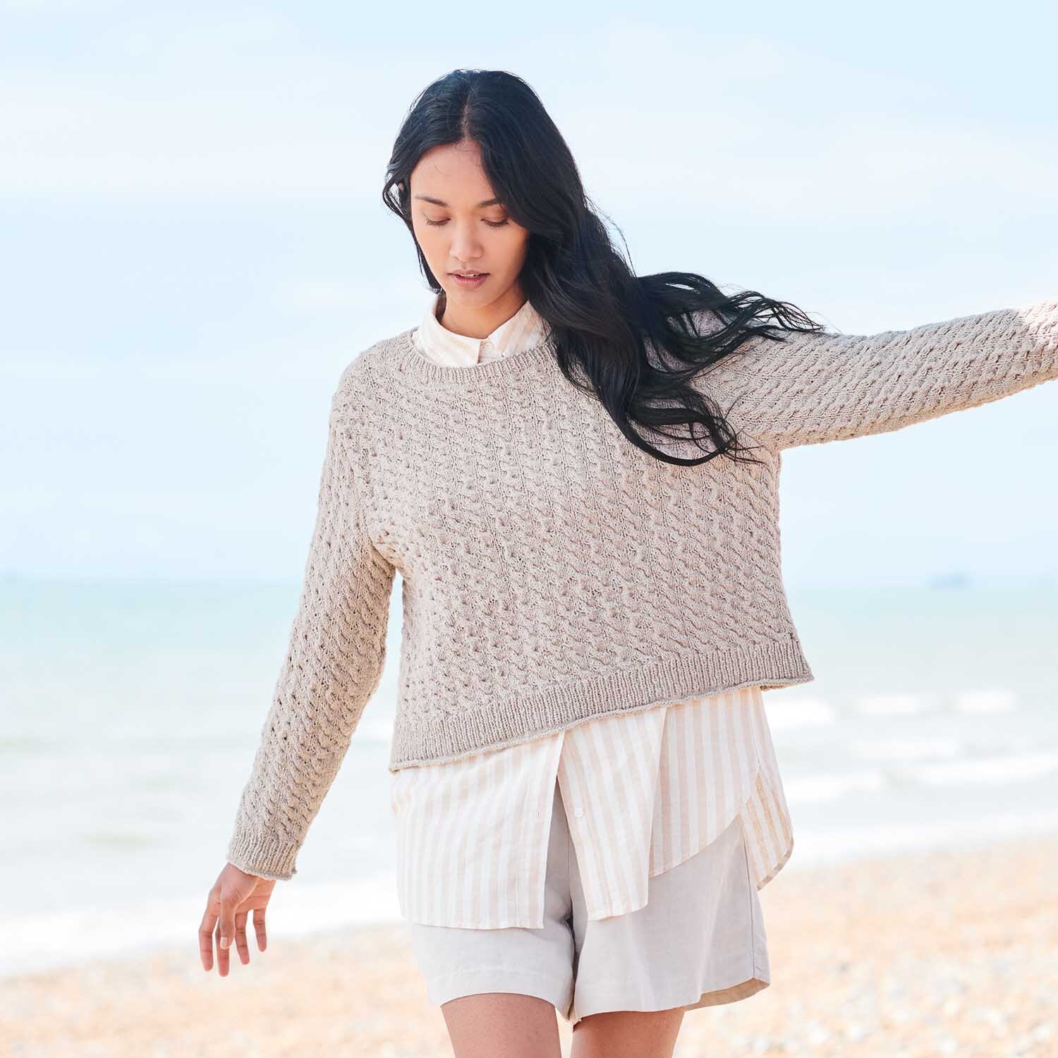 Strickanleitung Pullover aus Essentials Super Cotton dk: Frau am Strand in beigem Strickpullover, kombiniert mit heller Bluse und Shorts.