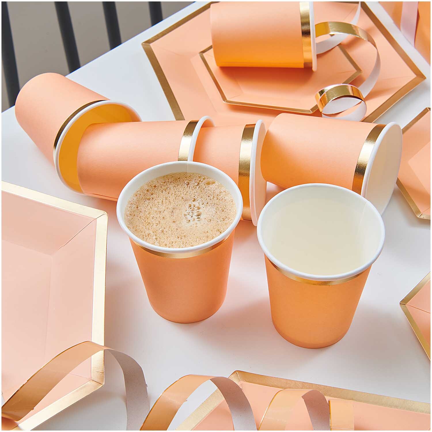 Pappbecher apricot-gold 250ml 10 Stück von Rico Design auf einem Tisch mit passenden Tellern und Bändern.