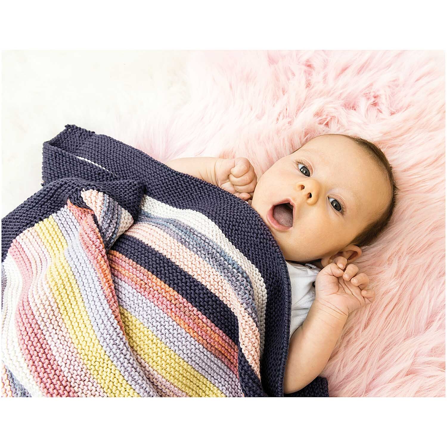 Rico Baby Cotton Soft dk: Ein Baby liegt auf einer rosa Decke, eingewickelt in eine bunte, gestrickte Decke.