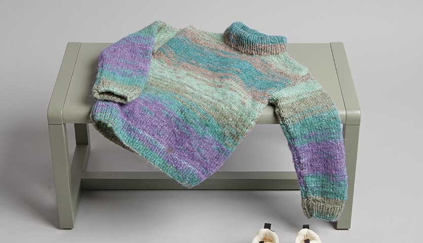 Strickanleitung Kinderpullover aus Creative Painted Power: Bunter Pullover auf grauer Bank, gestrickt in Pastelltönen.