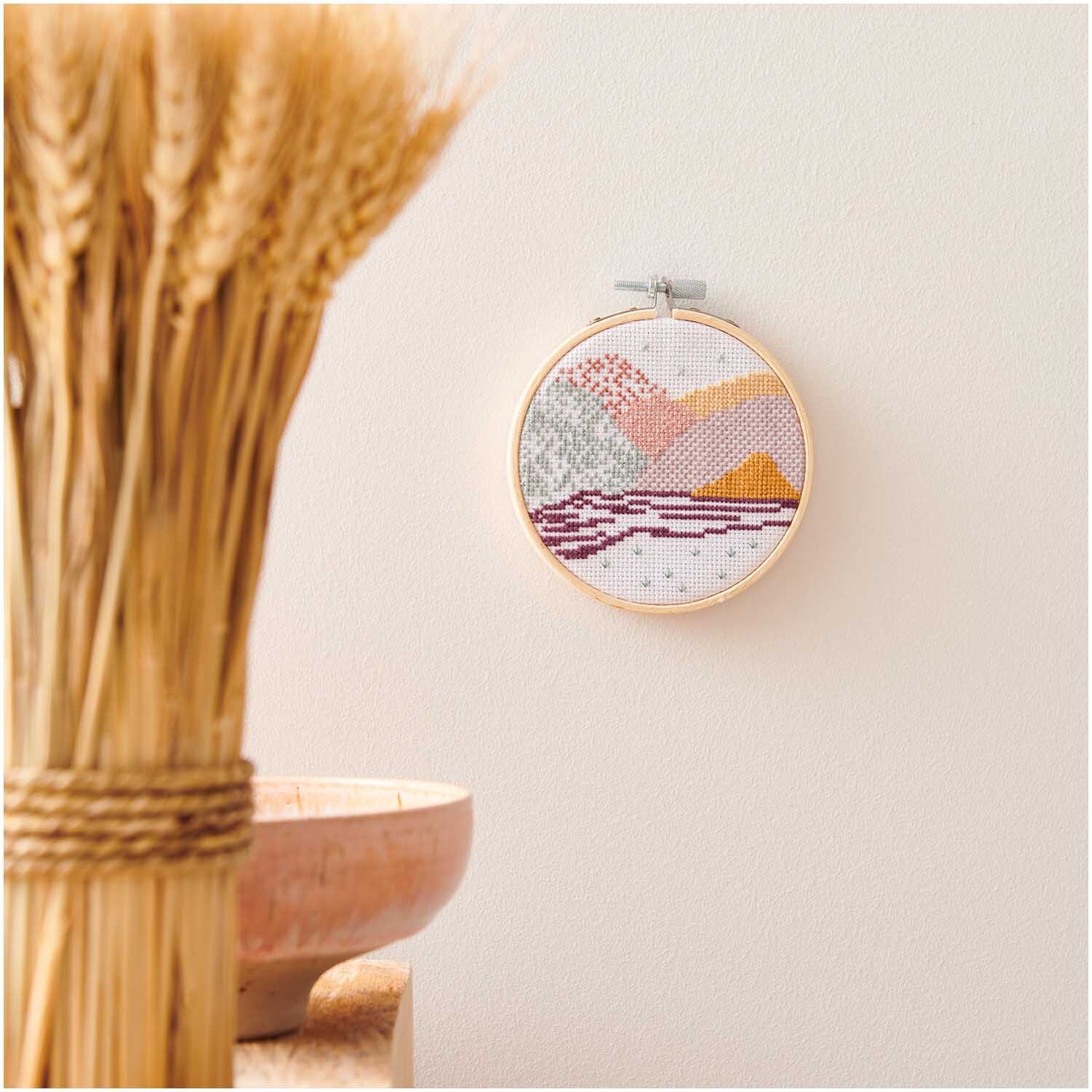 Stickpackung Mini Kit Landschaft Bild Ø 10cm an einer Wand, mit warmen Farbtönen und Holzrahmen.