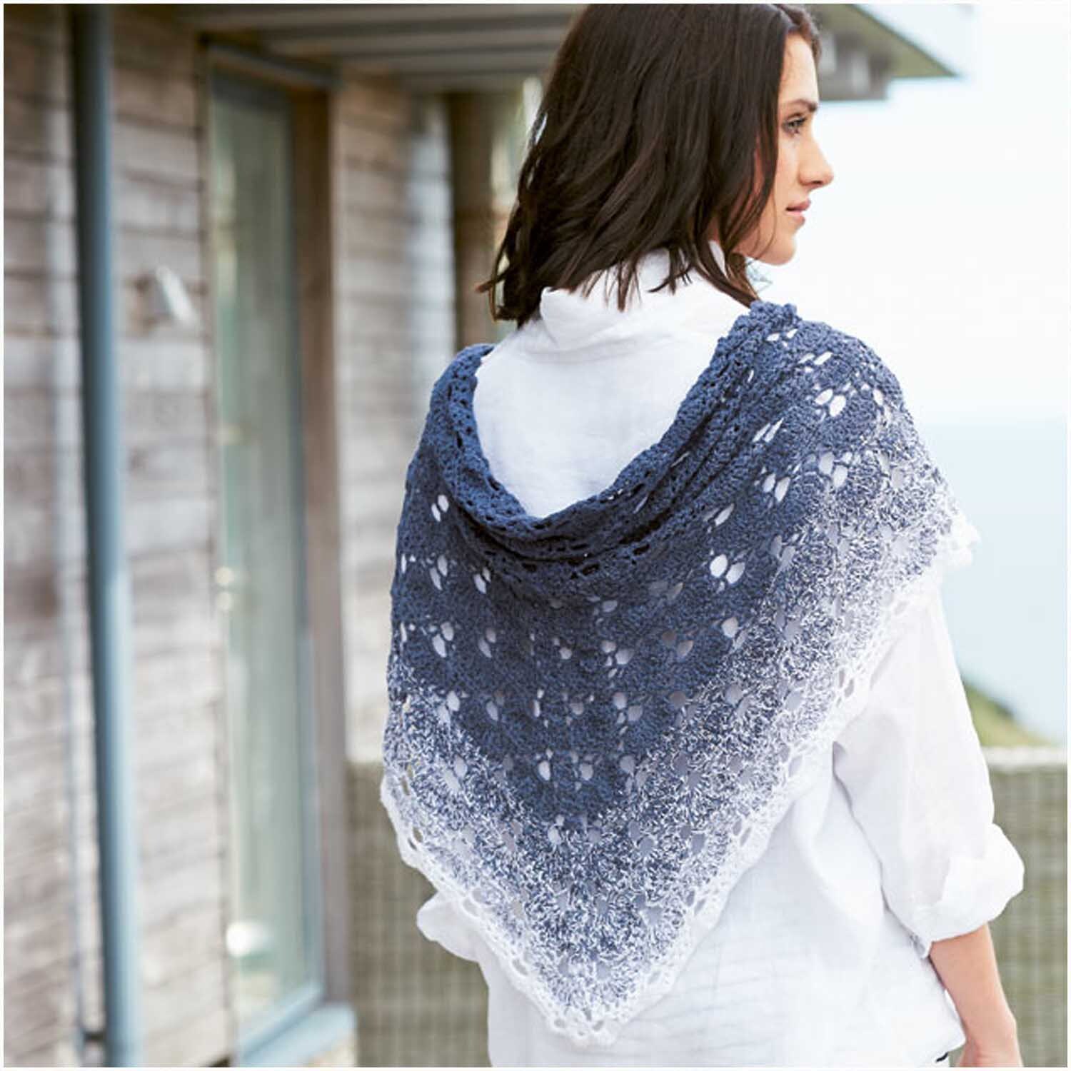 Creative Cotton dégradé von Rico Design: Frau trägt gestricktes Tuch in Blau-Weiß vor Holzhaus.