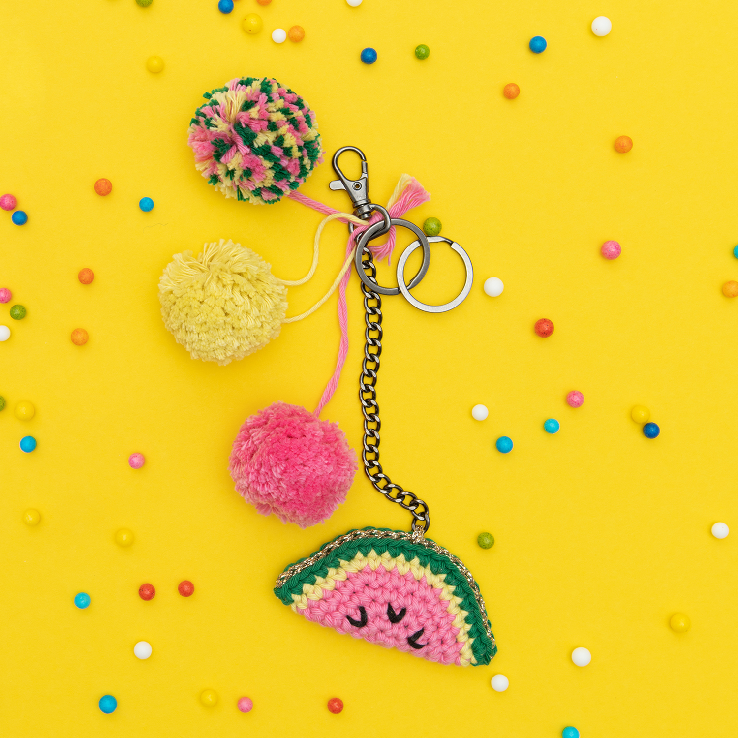 Häkelanleitung Obstanhänger: Bunter Schlüsselanhänger mit Pompons und gehäkeltem Wassermelonen-Design auf gelbem Hintergrund.