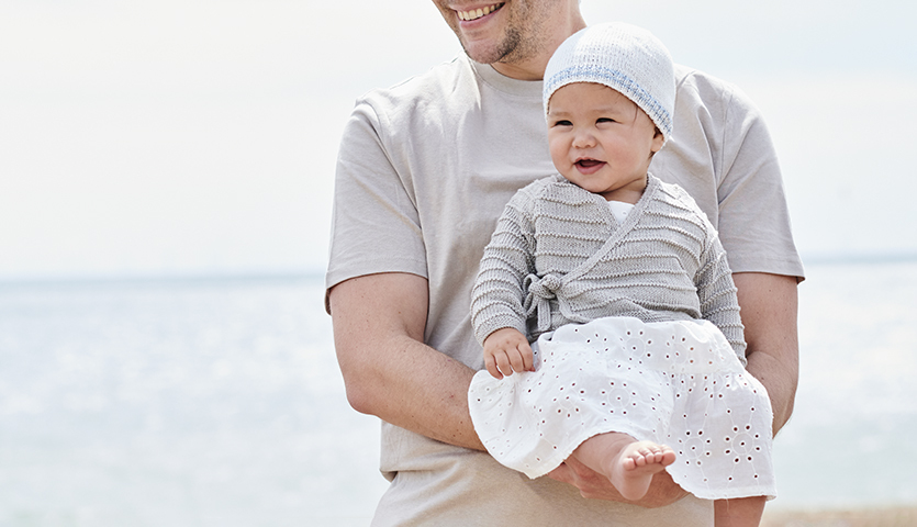 Strickanleitung Wickeljacke aus Baby Organic Cotton: Baby in grauer Wickeljacke und weißem Rock am Strand, gehalten von einem Erwachsenen.