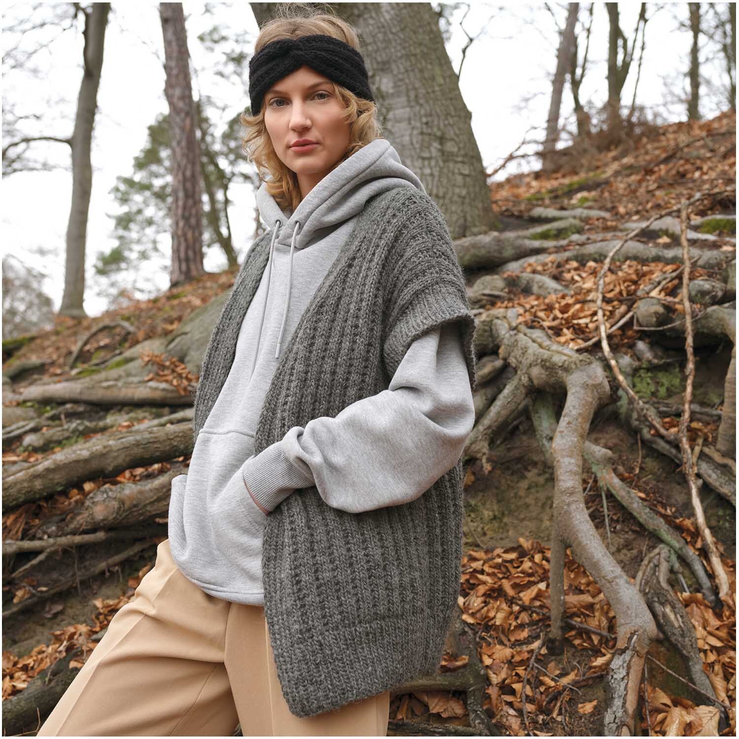 Essentials Organic Wool aran: Frau im Wald trägt graue Strickweste von Rico Design, kombiniert mit Kapuzenpullover und Stirnband.