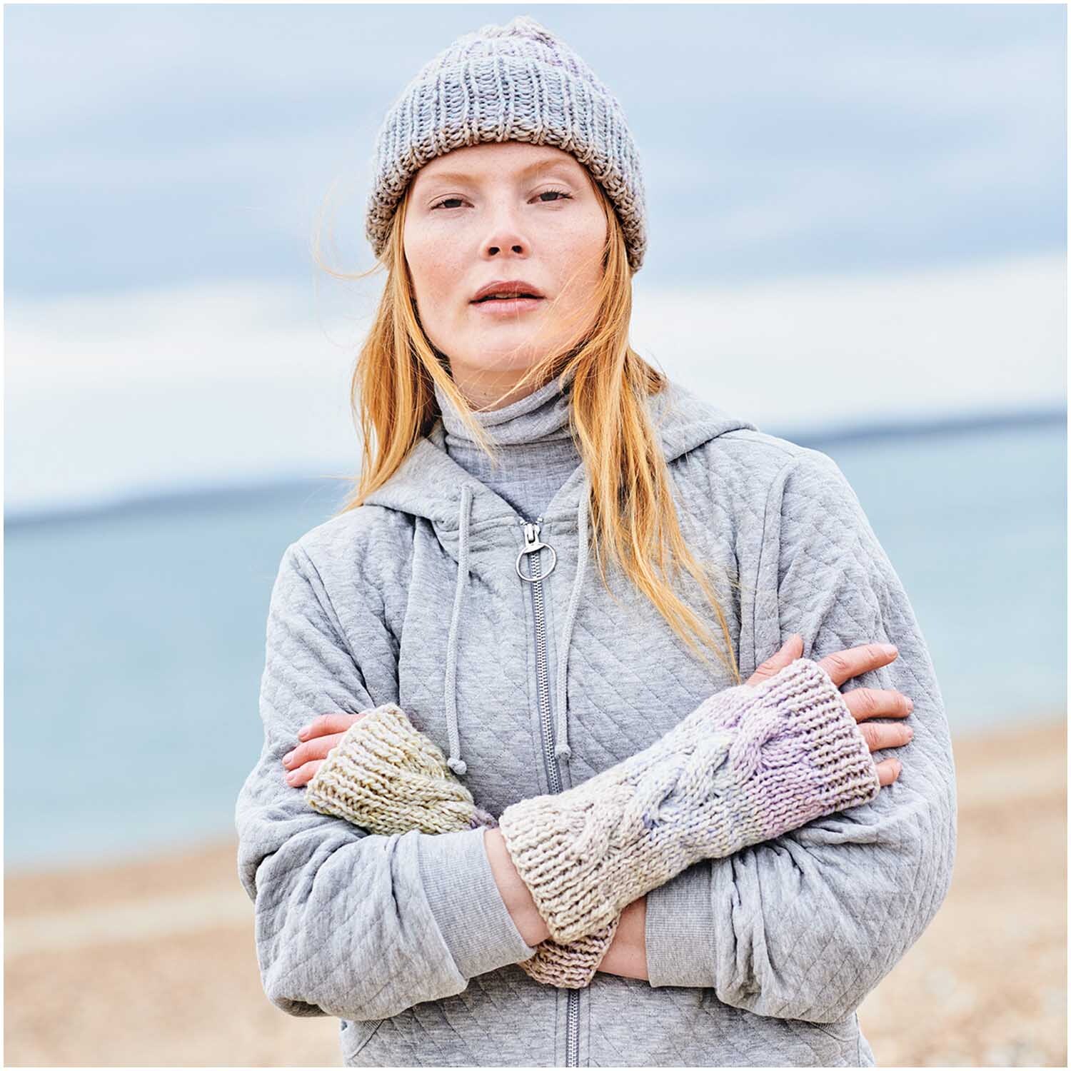 Creative Chic-Unique: Frau am Strand in grauem Outfit mit gestrickten Handschuhen und Mütze.
