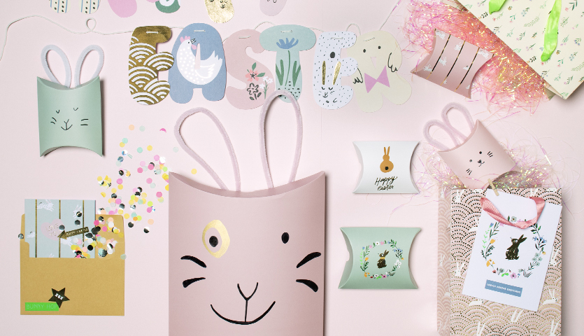 Bastelanleitung Geschenkschachtel Osterhase: Rosa Schachtel mit Hasenmotiv, umgeben von bunten Dekorationen und Karten.