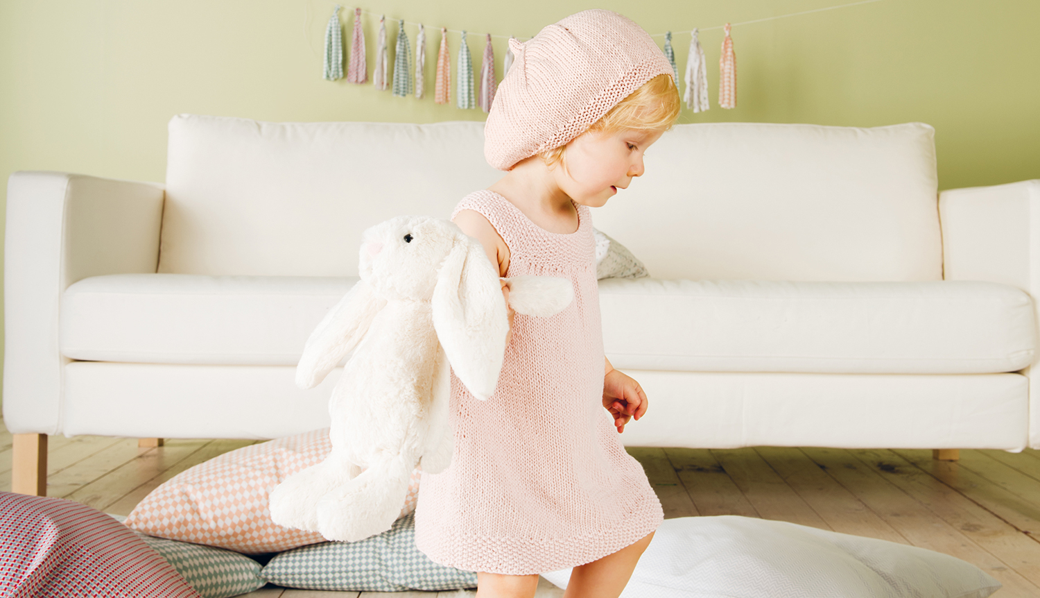 Strickanleitung Rico Baby 19: Kleinkind in rosa Kleid und Mütze hält ein weißes Kuscheltier vor einem Sofa.