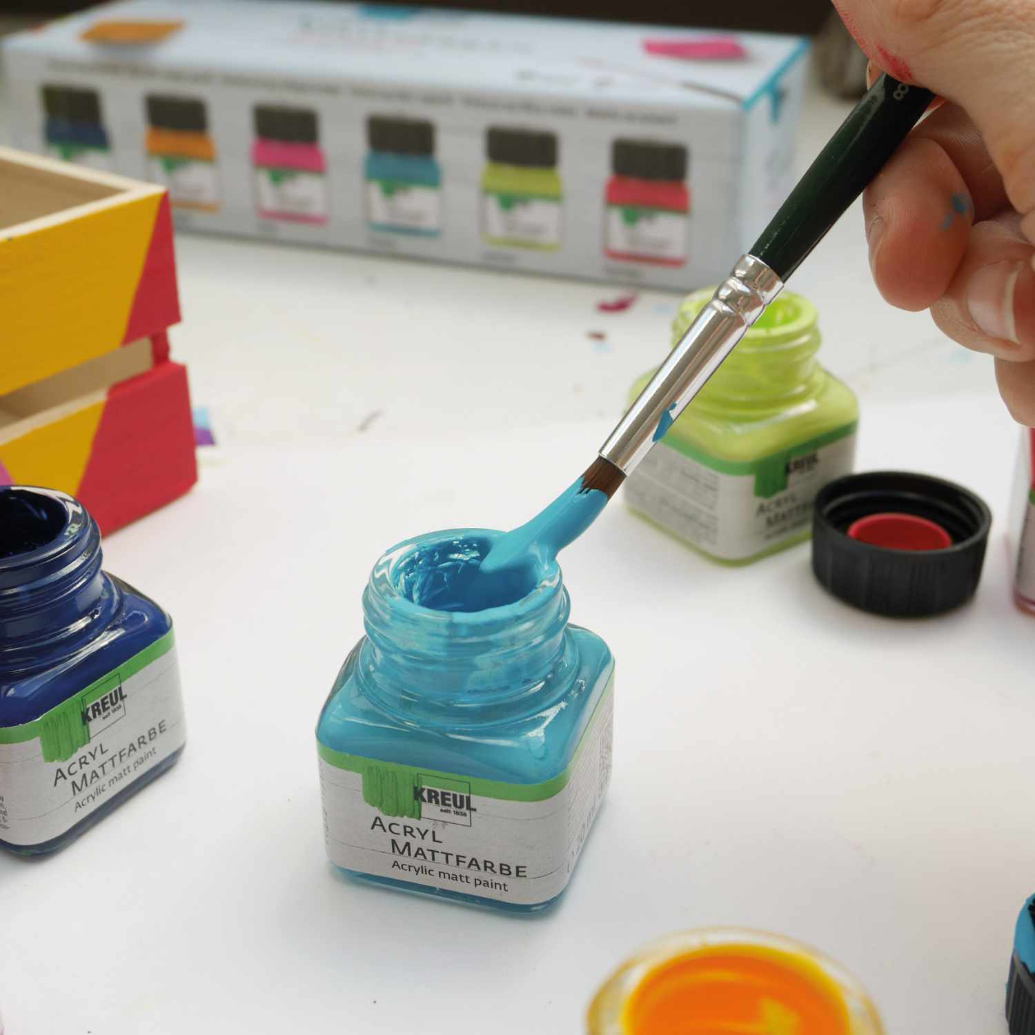 Acryl Mattfarben Set Color Living 6x20ml: Hand taucht Pinsel in blaue Farbe von KREUL, umgeben von weiteren Farbtöpfen.