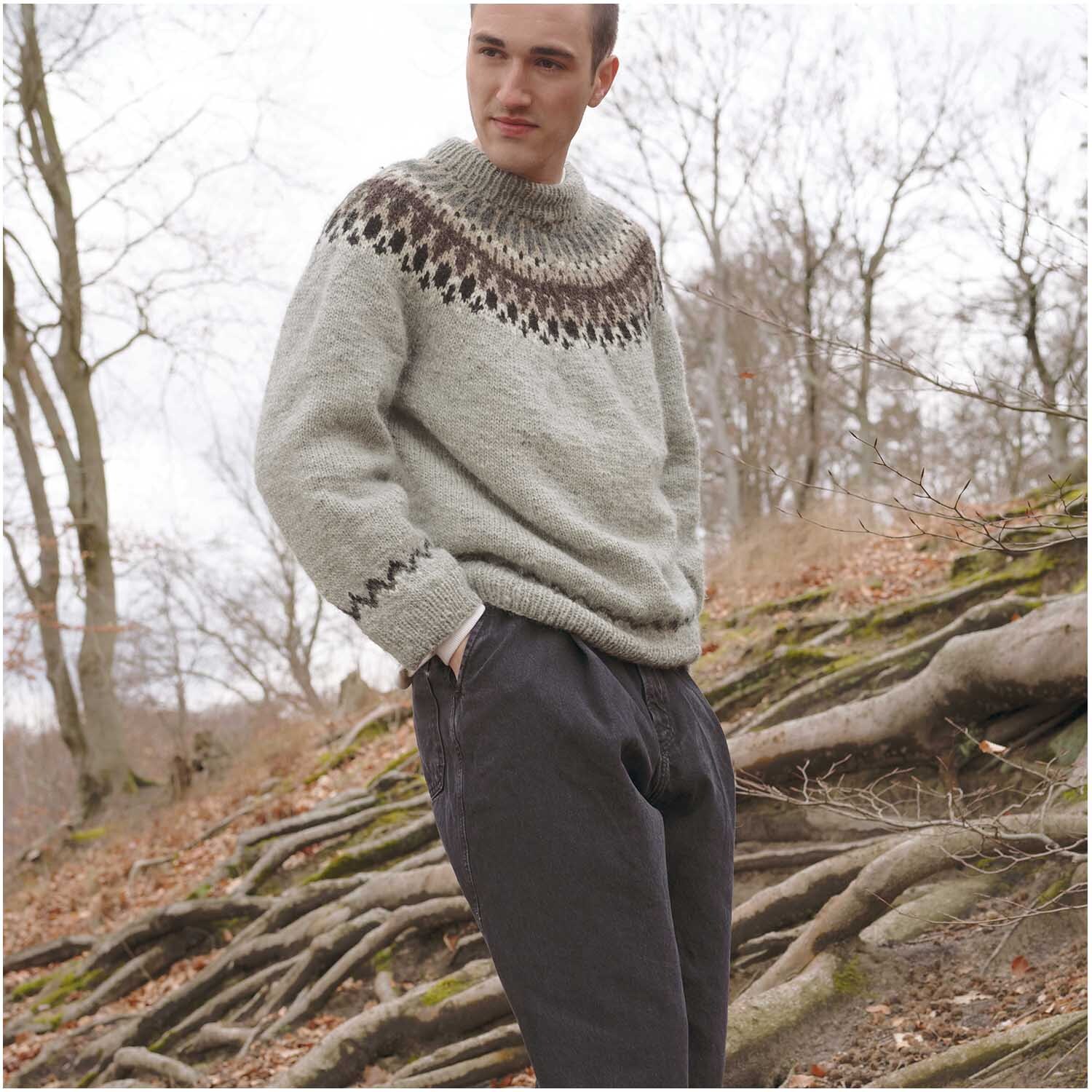 Essentials Organic Wool aran: Person in grauem Strickpullover mit Muster, stehend im Wald. Ideal für kühle Tage.