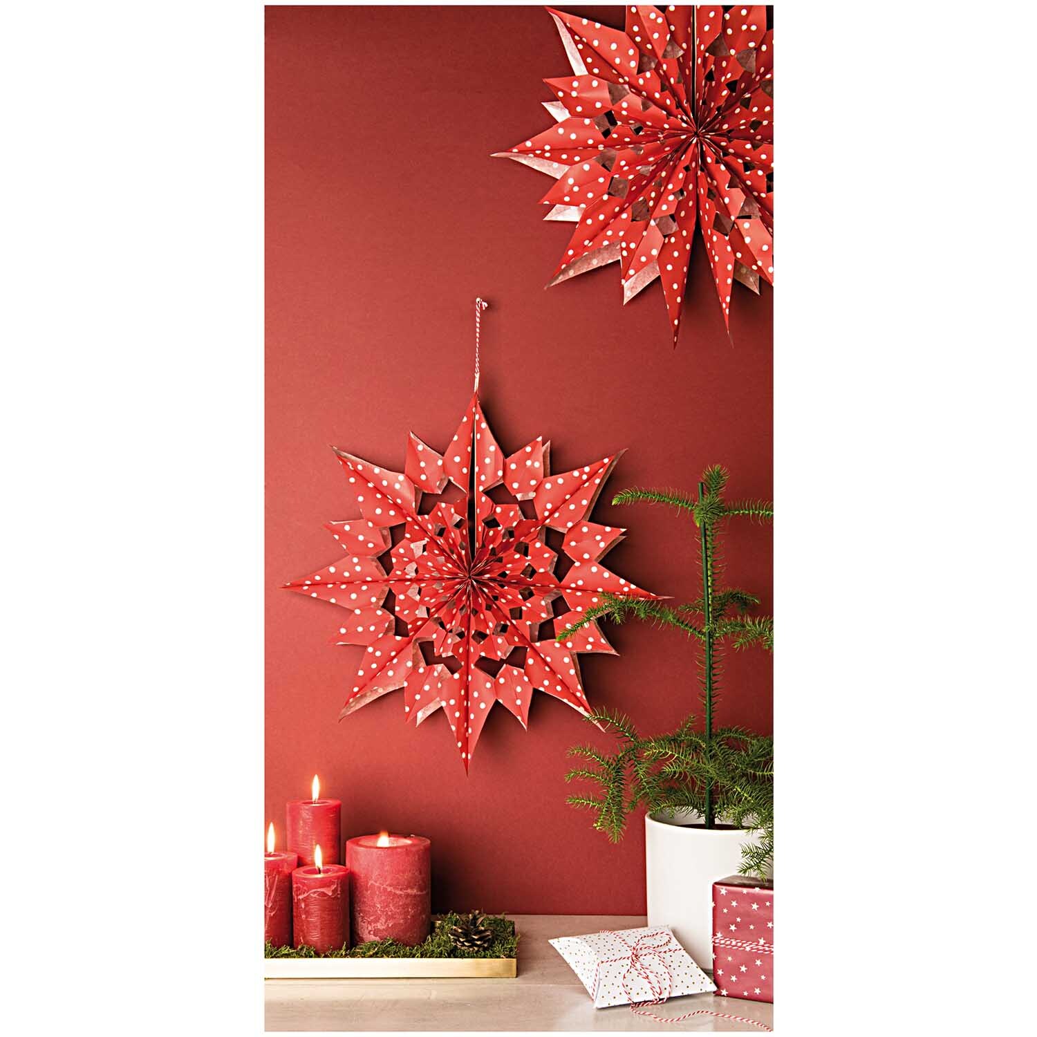 Paper Poetry Bastelset Papiertüten-Sterne Punkte groß: Zwei rote, gepunktete Papiersterne hängen an einer roten Wand, umgeben von Kerzen und Geschenken.