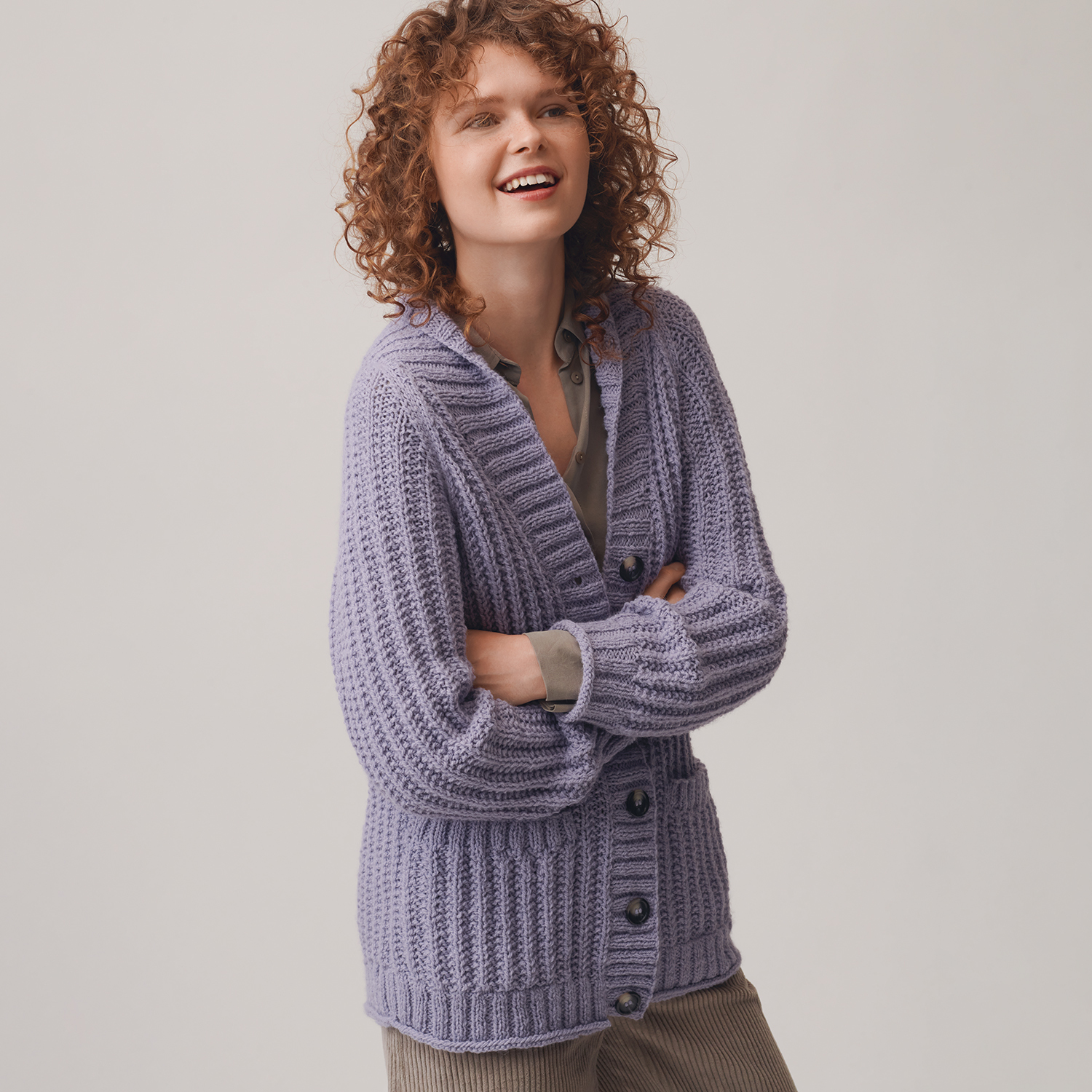 Strickanleitung Damenjacke aus Creative Soft Wool aran: Frau trägt eine lila Strickjacke mit Knöpfen, lässig über einem grauen Oberteil und beigen Hosen.