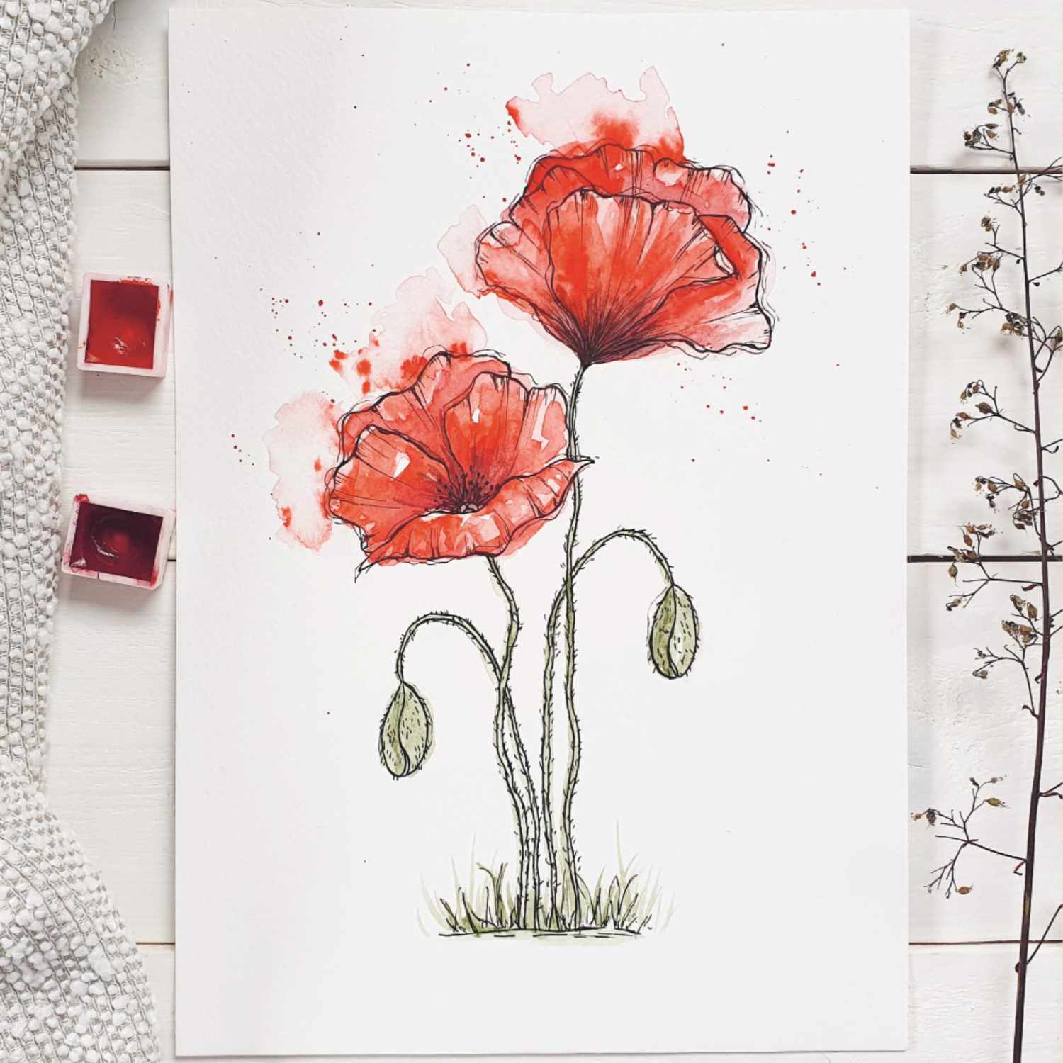 Anleitung Watercolor Mohnblume: Zwei rote Mohnblumen in Aquarelltechnik auf weißem Papier, umgeben von Malutensilien.