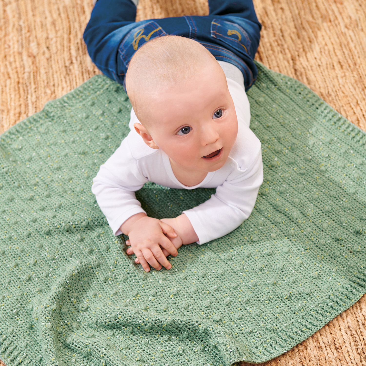 Häkelanleitung Babydecke aus Baby Dream Tweed dk: Ein Baby liegt auf einer grünen Decke auf einem Teppich.