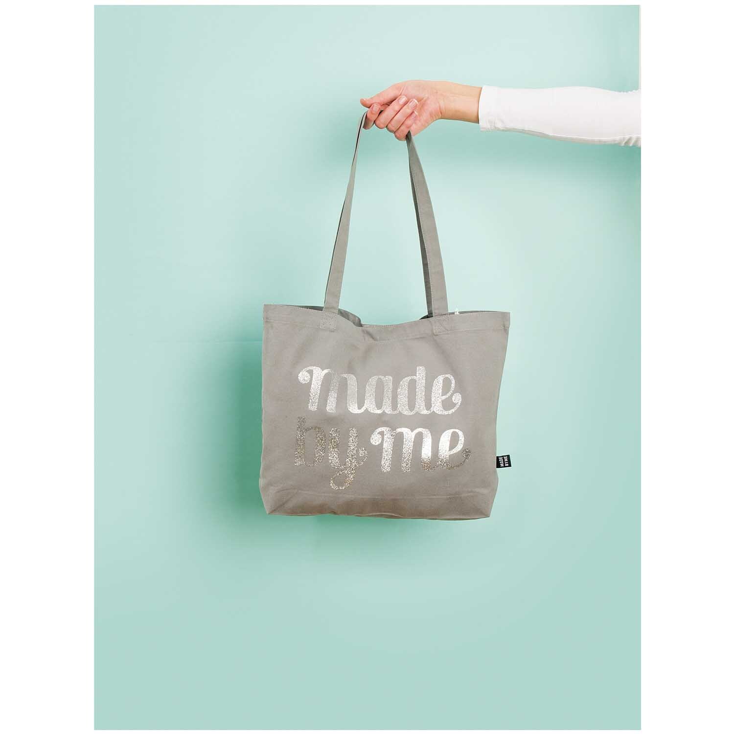 Tasche mit langen Henkeln, graue Farbe, von Rico Design, gehalten von einer Hand vor mintgrünem Hintergrund.