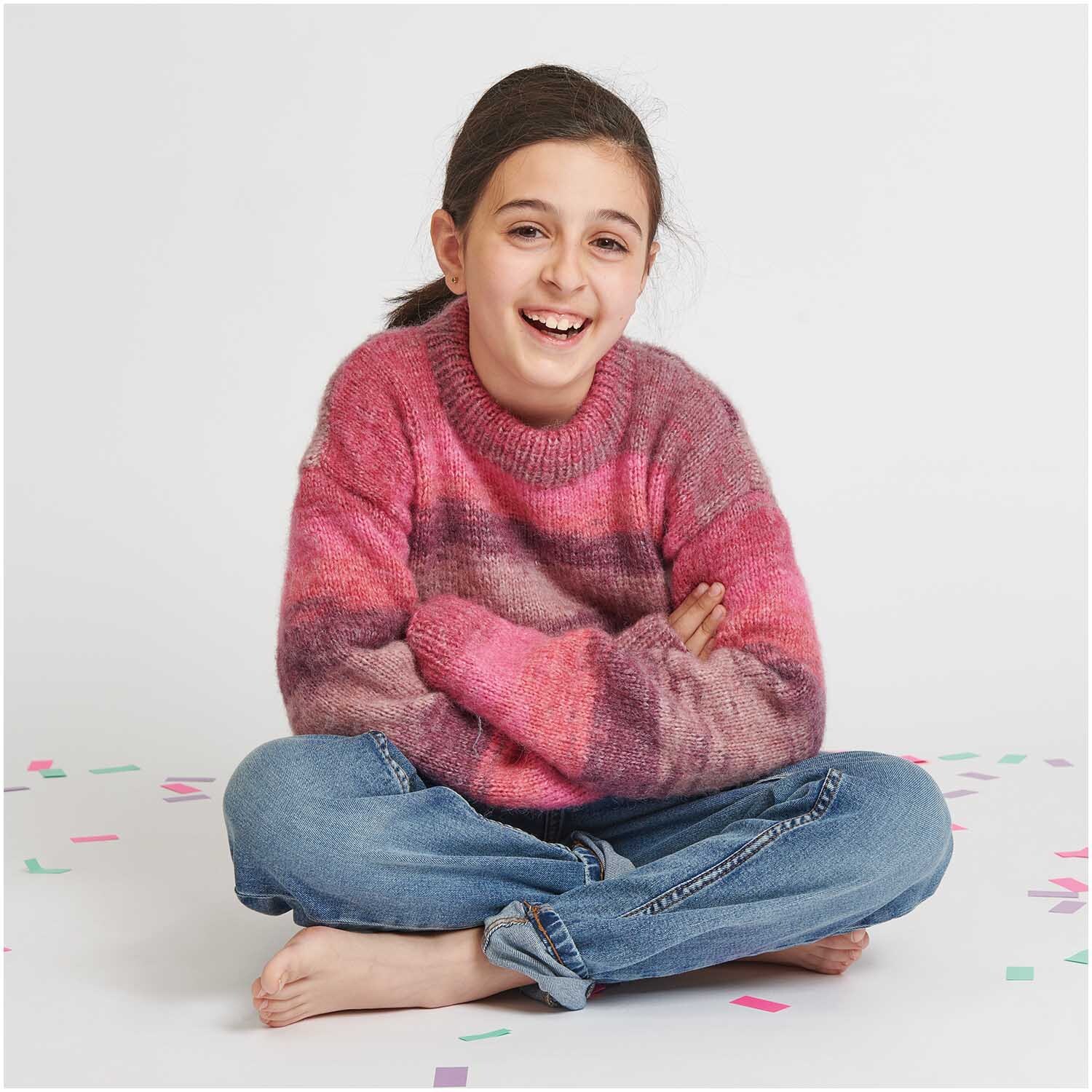 Creative Painted Power: Mädchen in einem gestrickten Pullover in Beerenfarben von Rico Design, sitzt lachend auf dem Boden.