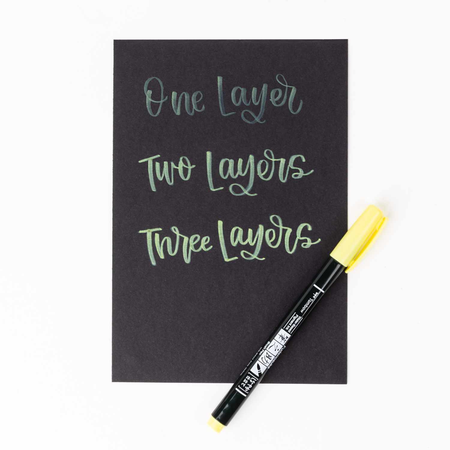 Fudenosuke Brush Pen Pastel von Tombow auf schwarzem Papier, zeigt Schrift in verschiedenen Schichten.