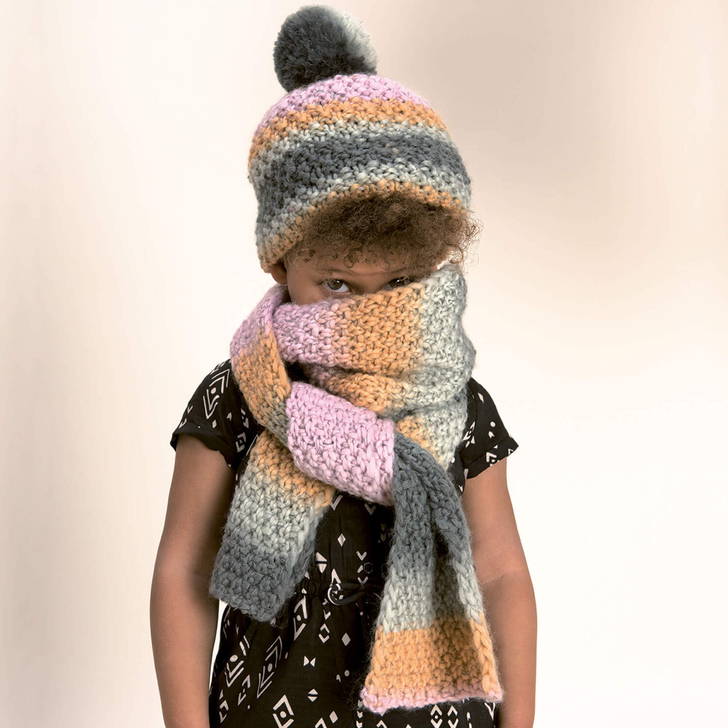 Strickanleitung Kinderschal und Mütze aus Glühwürmchen Garn: Kind trägt gestreifte Mütze und Schal in Pastellfarben, warm und stilvoll für kalte Tage.