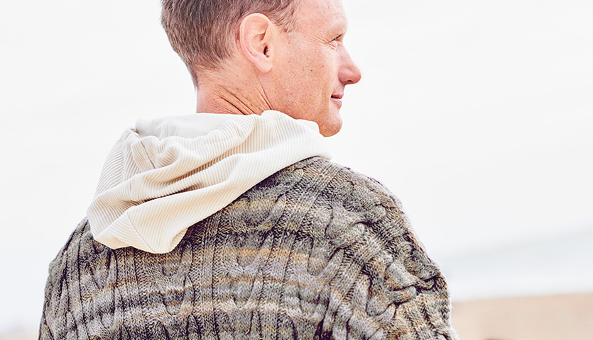Strickanleitung Pullover aus Creative Melange aran Wonderball: Mann in gemustertem Strickpullover mit Kapuze am Strand.
