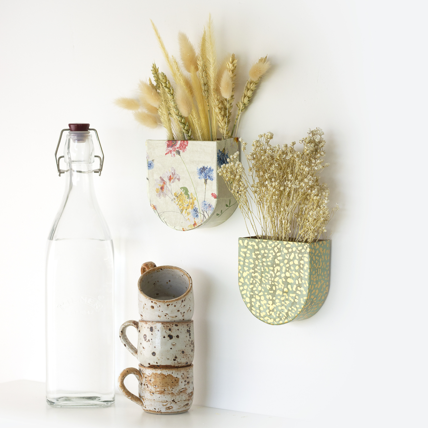 Bastelset Kreativer Moment Wandhalter: Zwei dekorative Wandhalter mit Trockenblumen neben einer Glasflasche und gestapelten Tassen.