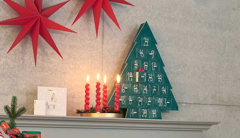Anleitung Holz-Adventskalender in Shabby-Chic-Look: Grüner Baum mit roten Zahlen, dekoriert mit Kerzen und roten Sternen auf einem Regal.