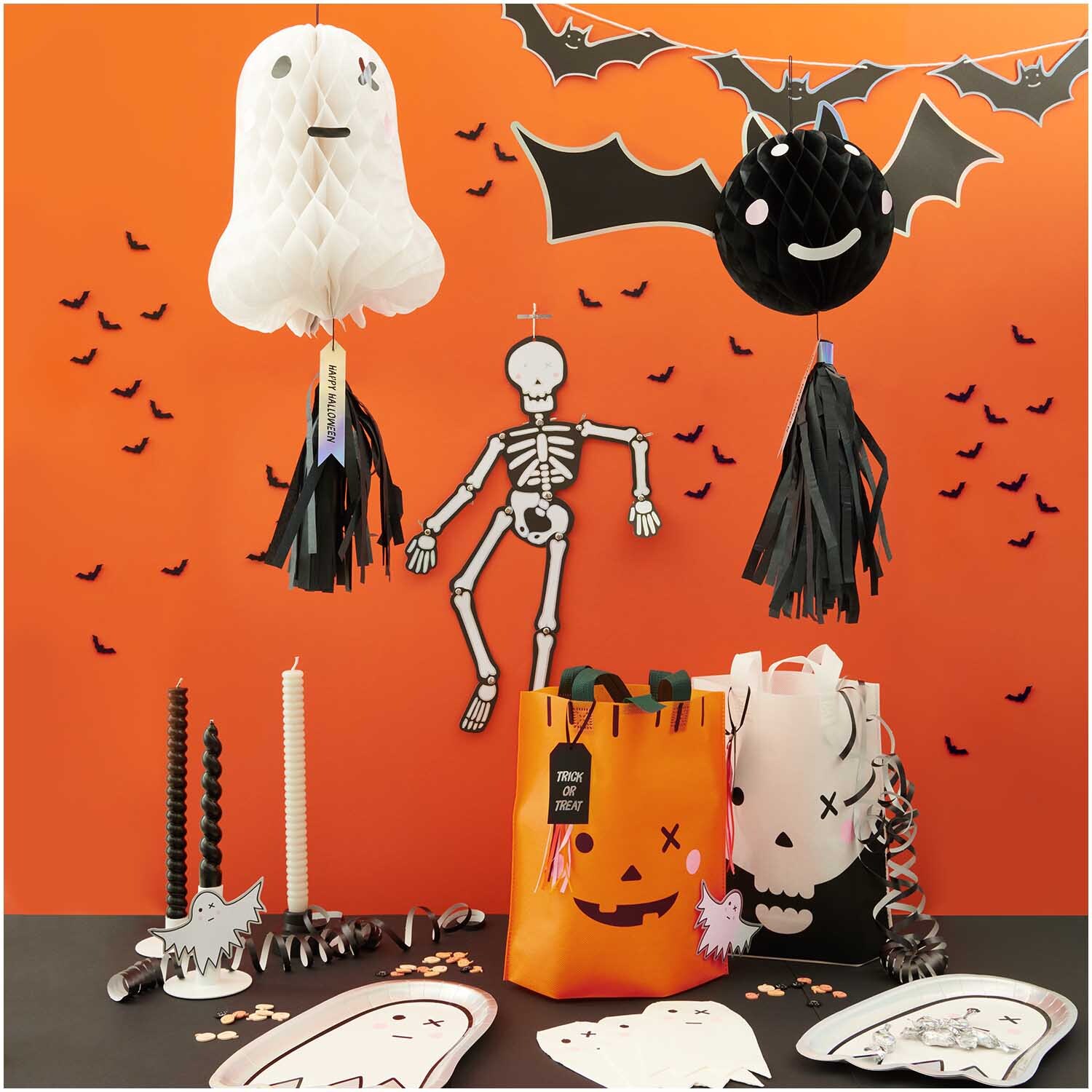 Paper Poetry Geschenktüte Kürbis 28x30cm vor Halloween-Dekoration mit Skelett, Fledermaus und Geistern auf orangefarbenem Hintergrund.