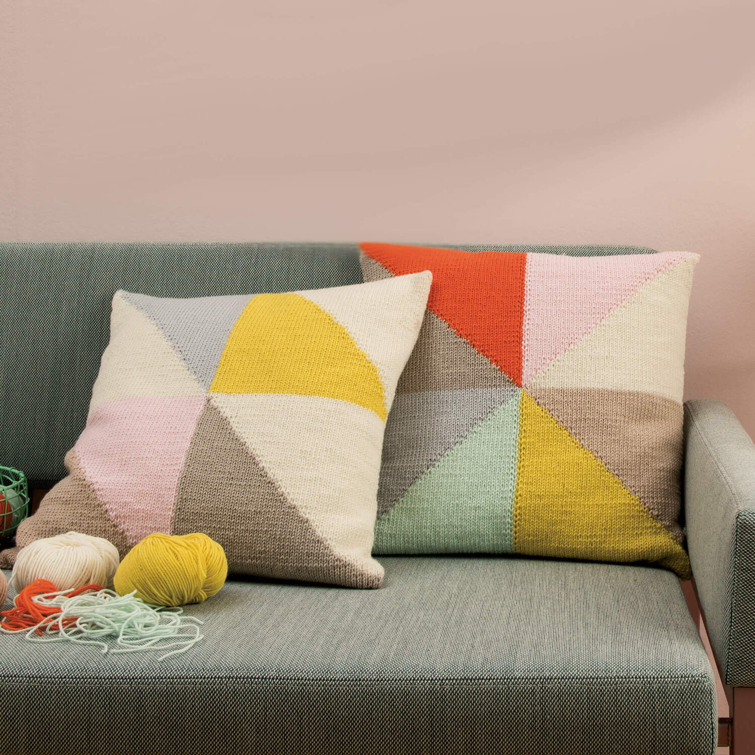 Strickanleitung Kissenhülle aus Essentials Merino: Zwei bunte Kissen auf einem Sofa, gestrickt in geometrischen Mustern mit Garnknäueln im Vordergrund.