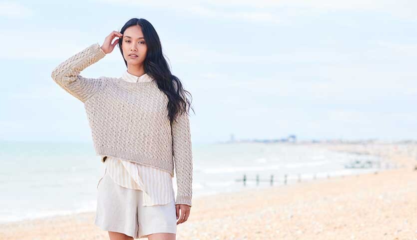 Strickanleitung Pullover aus Essentials Super Cotton dk: Frau am Strand in beigem Pullover und weißem Hemd.