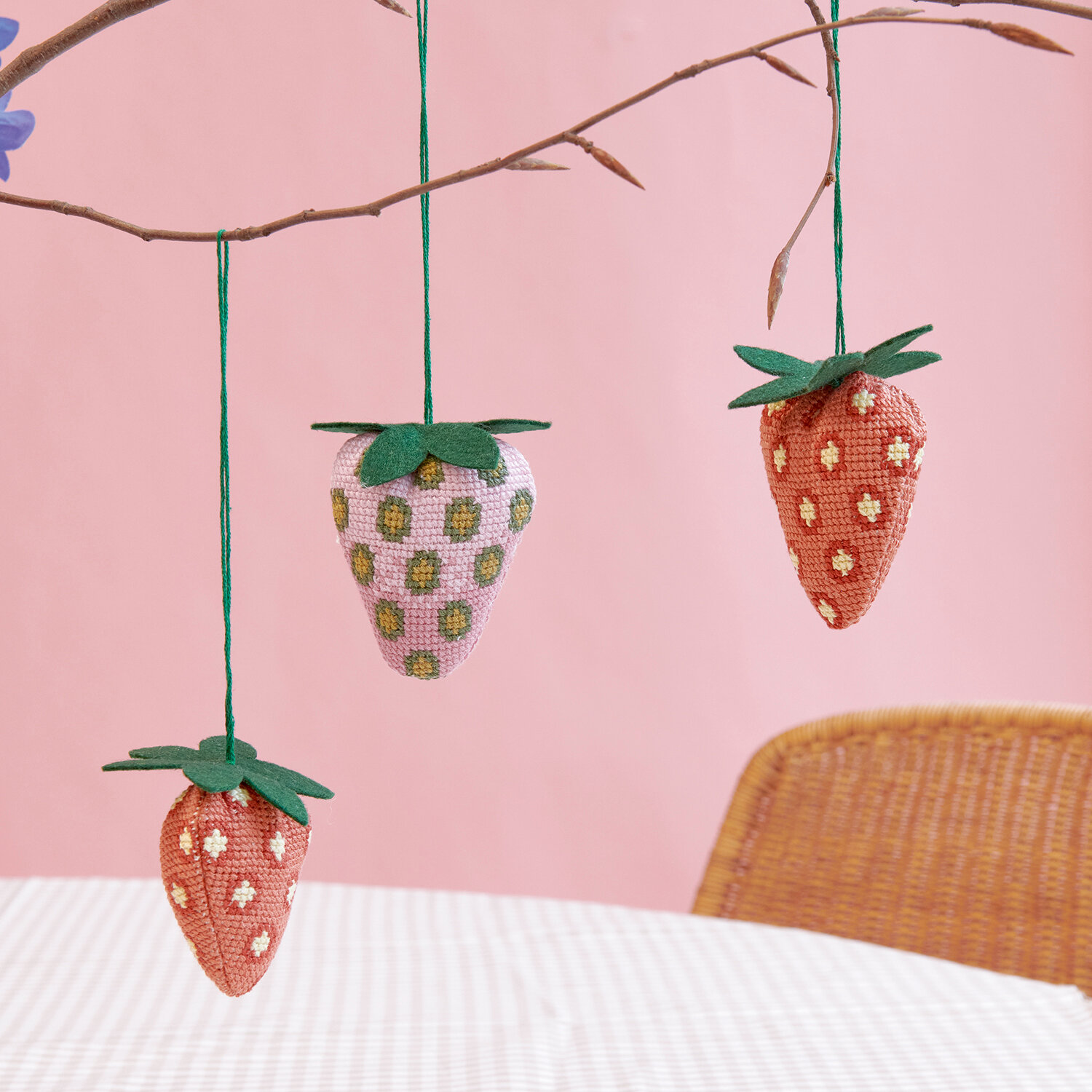 Stickanleitung Erdbeer-Anhänger mit gehäkelten Deko-Erdbeeren in Pastell- und Rot-Tönen, hängend an einem Ast vor rosa Wand, perfektes DIY-Nähprojekt