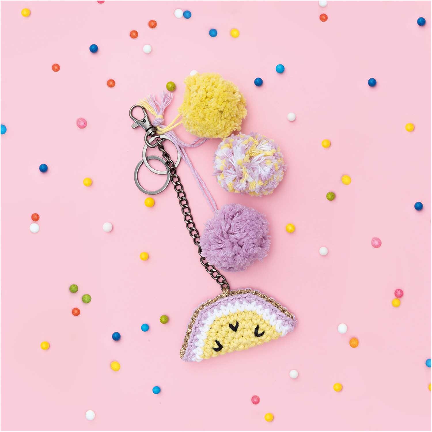Creative Cotton aran Schlüsselanhänger mit bunten Pompons und gehäkeltem Smiley auf rosa Hintergrund.