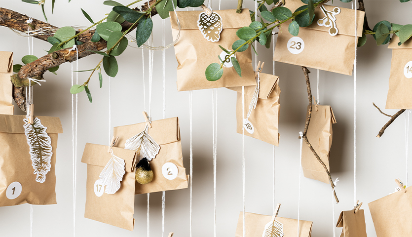 Bastelanleitung Papiertüten-Adventskalender: Braune Papiertüten an Zweigen, dekoriert mit Blättern und Zahlen, ideal für kreative Adventsdekoration.