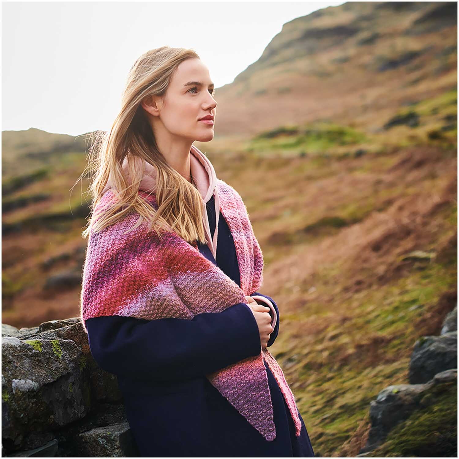 Strickset Dreieckstuch Modell 14: Frau trägt ein rosa-rot gemustertes Tuch in einer natürlichen Landschaft.