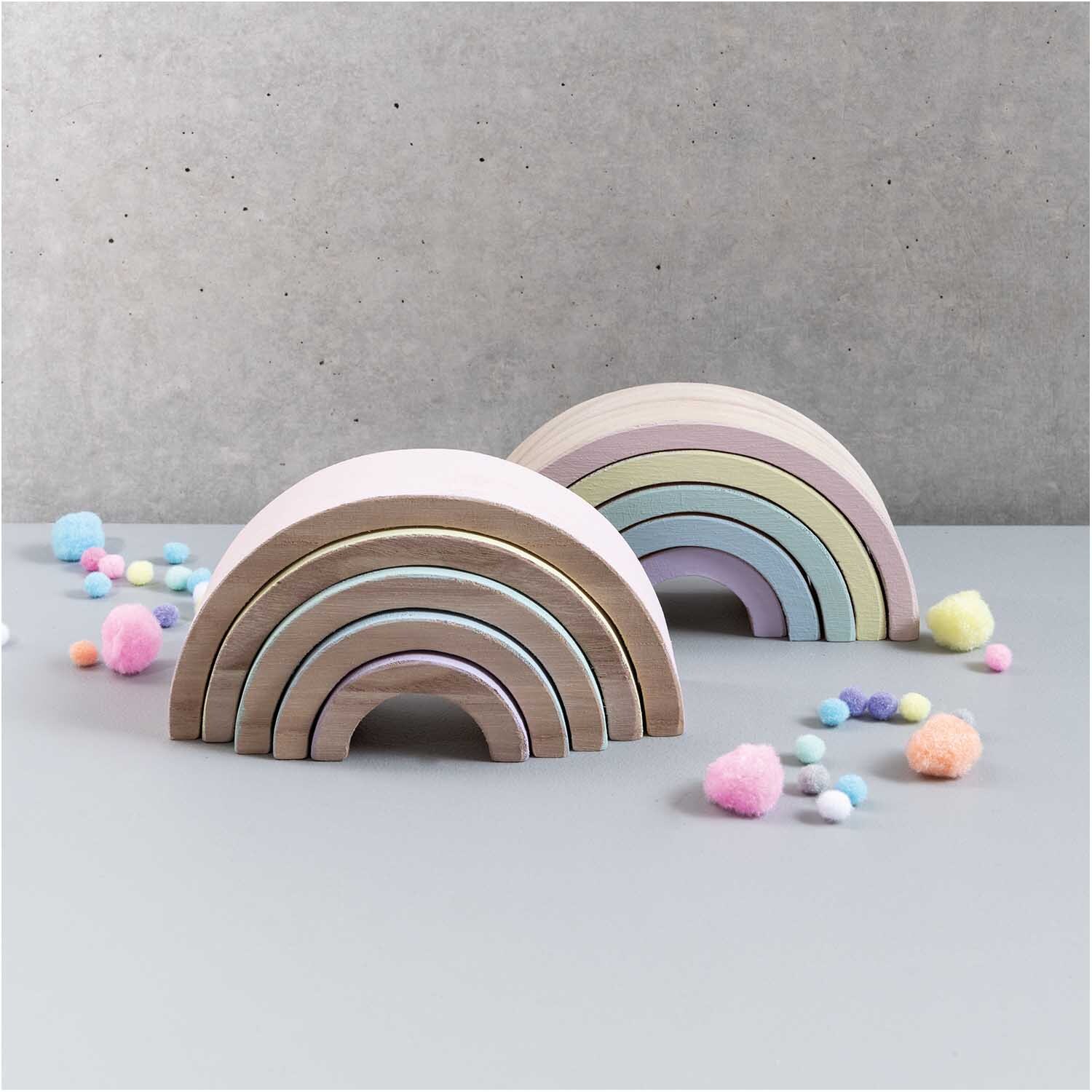 Holz-Regenbogen 18x9x5cm 5teilig: Zwei pastellfarbene Holz-Regenbögen von Rico Design auf grauem Hintergrund, umgeben von bunten Pompons.