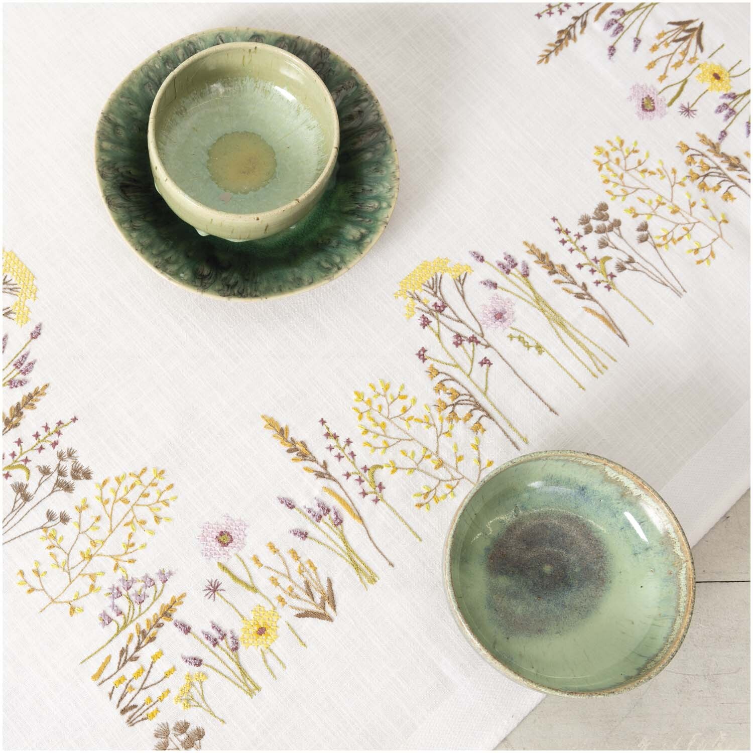 Stickpackung Tischdecke Kräuterwiese 90x90cm mit floralen Mustern und zwei grünen Keramikschalen, ideal für stilvolle Tischdekoration.