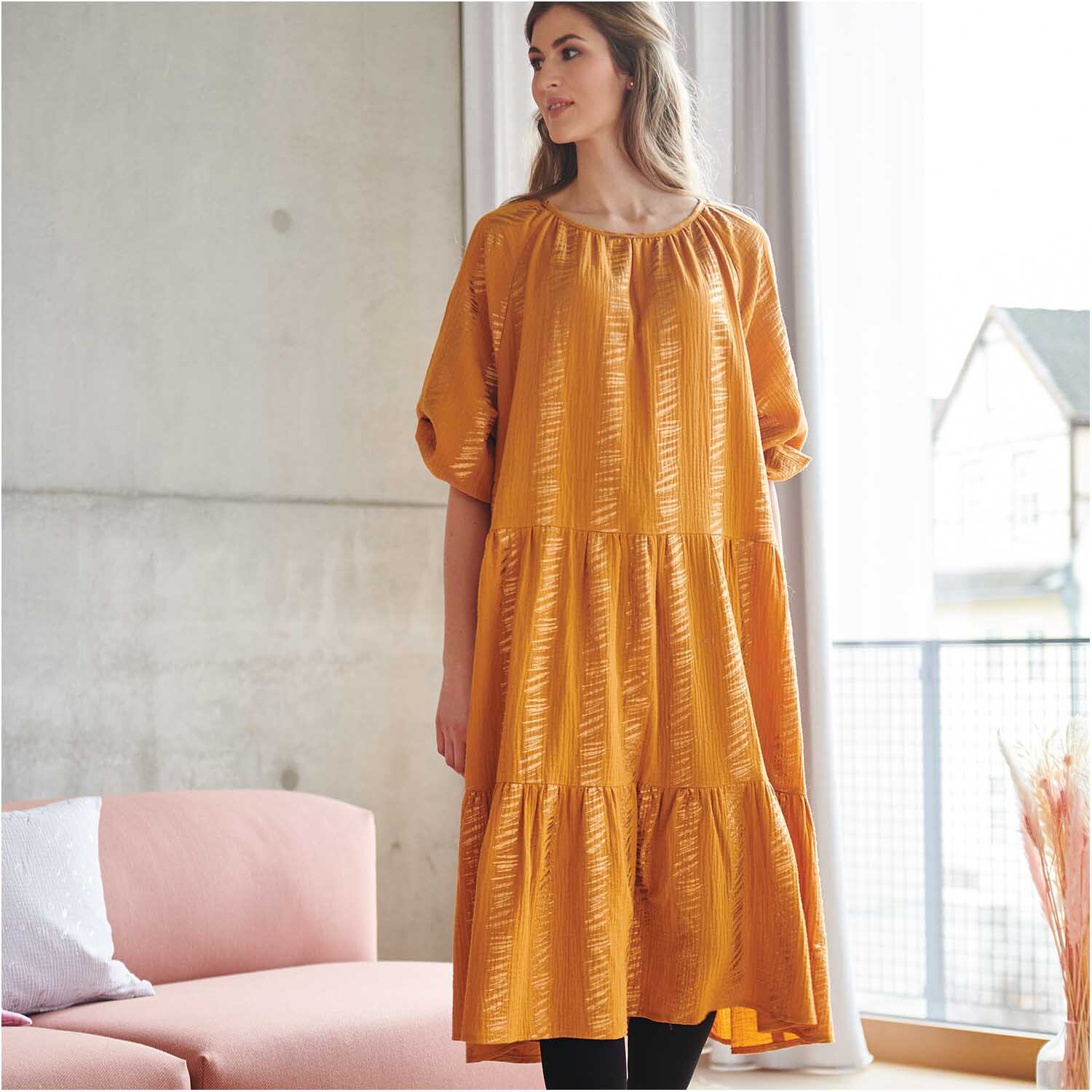 Stoffabschnitt Krinkelmusselin senf 50x130cm Hot Foil: Frau in senffarbenem Kleid von Rico Design steht in modernem Wohnzimmer.