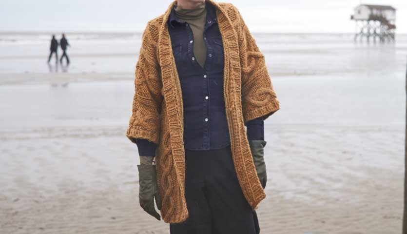 Strickanleitung Zopfjacke aus Creative Twist super chunky: Frau am Strand in grob gestrickter, brauner Jacke, kombiniert mit dunkler Kleidung.