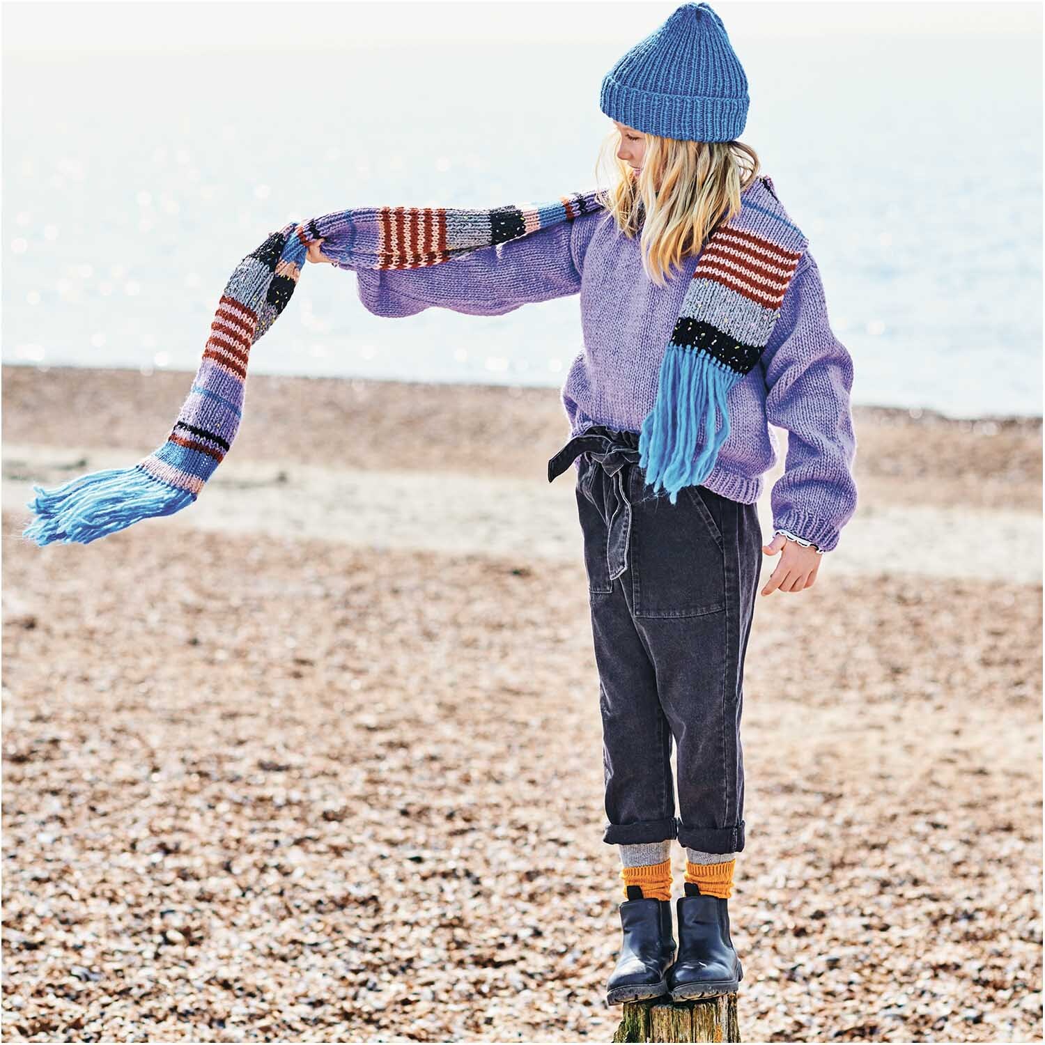 Essentials Super aran: Kind in lila Pullover und Mütze am Strand, trägt gestreiften Schal.