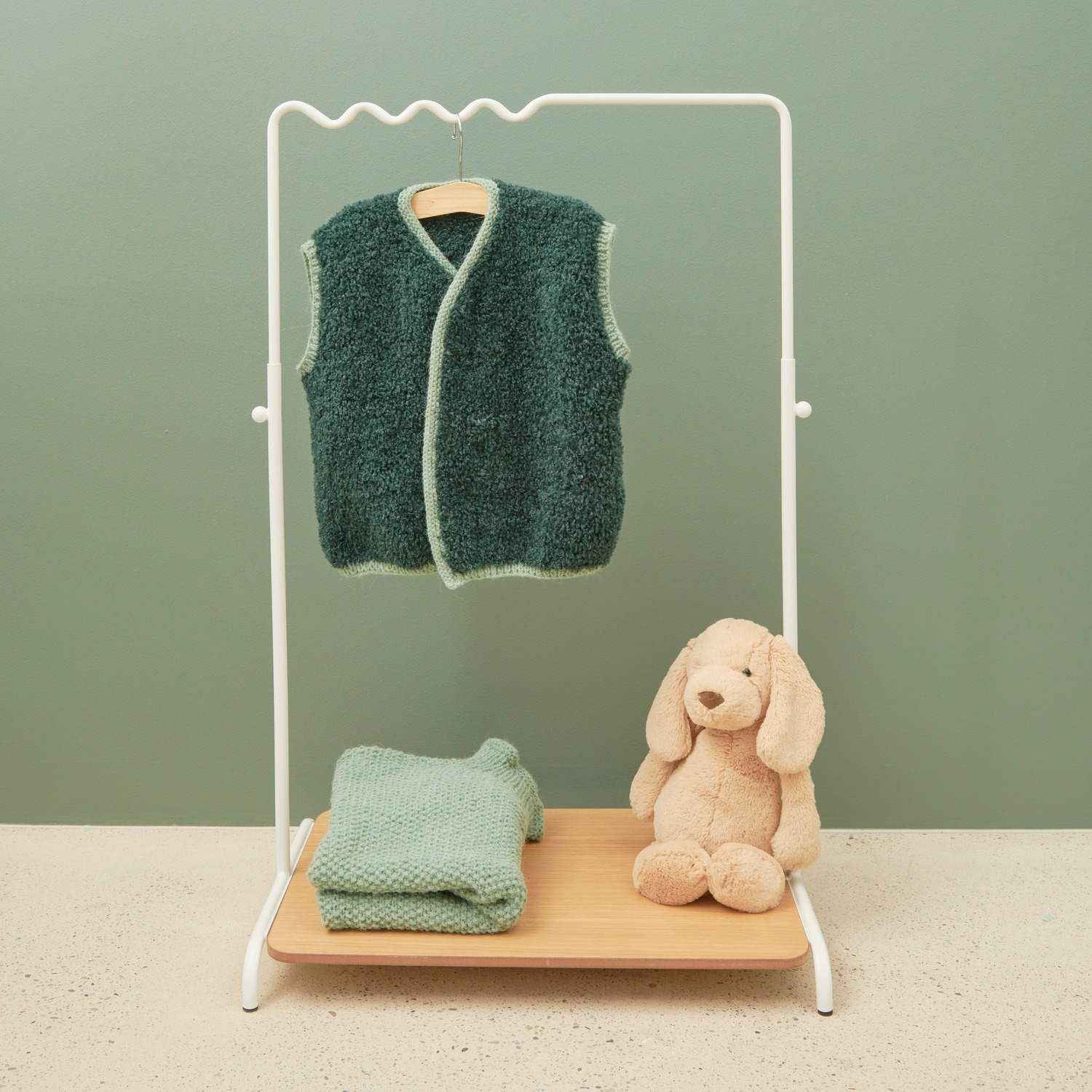 Rico Kids 13: Kinderweste und Pullover in Petrol auf weißem Kleiderständer mit Plüschhase.