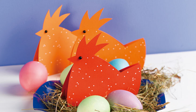 Bastelanleitung Hühner aus Papier: Drei orangefarbene Papierhühner mit bunten Eiern auf Heu.