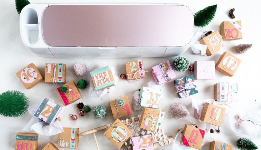 Bastelanleitung Adventskalender plotten mit dem Cricut Maker: Kreative Boxen in Pastellfarben, umgeben von Tannenzweigen und Deko.