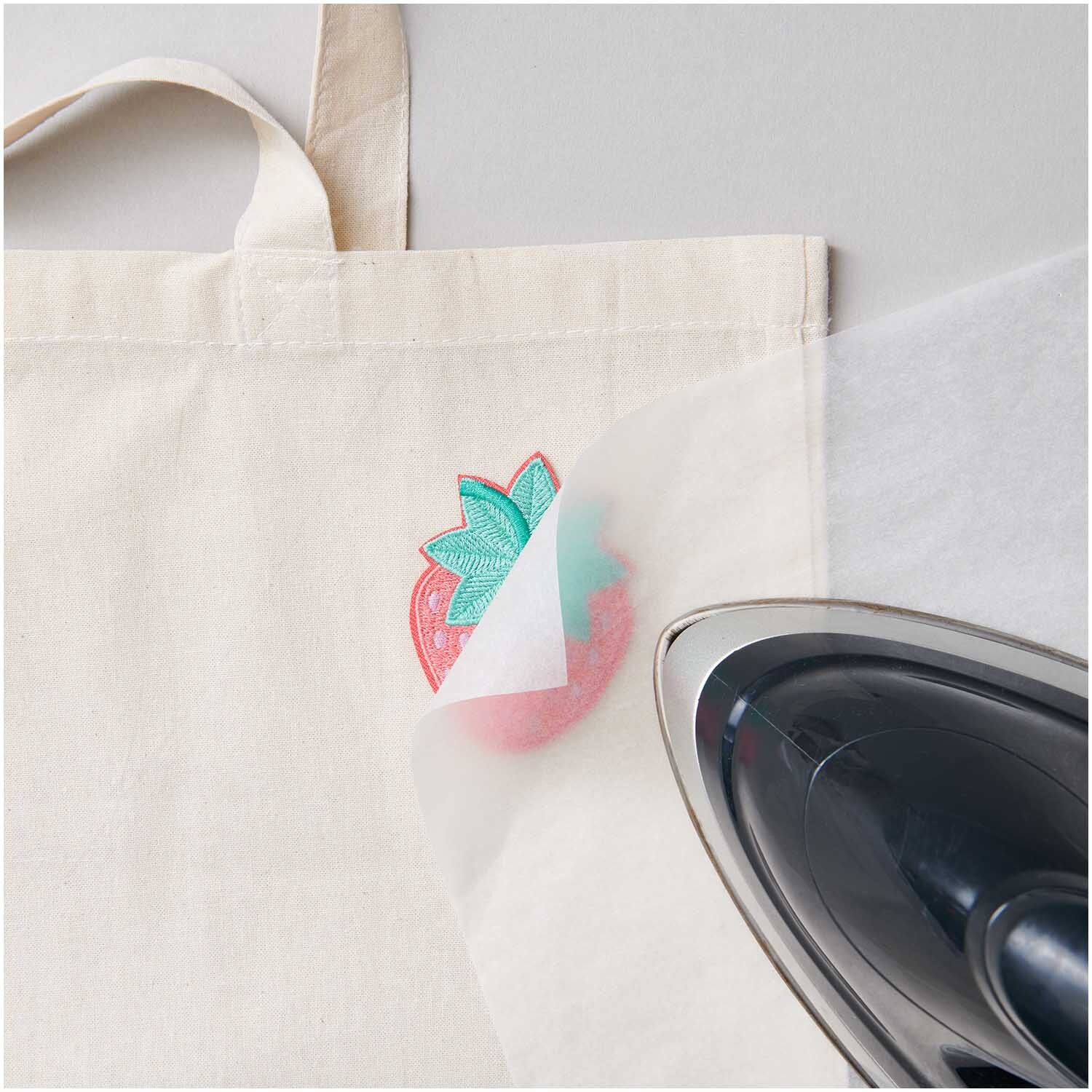 Patch Erdbeere zum Aufbügeln 5x6,8cm auf einer Tasche, mit Bügeleisen und Schutzpapier. Ideal für kreative Designs.