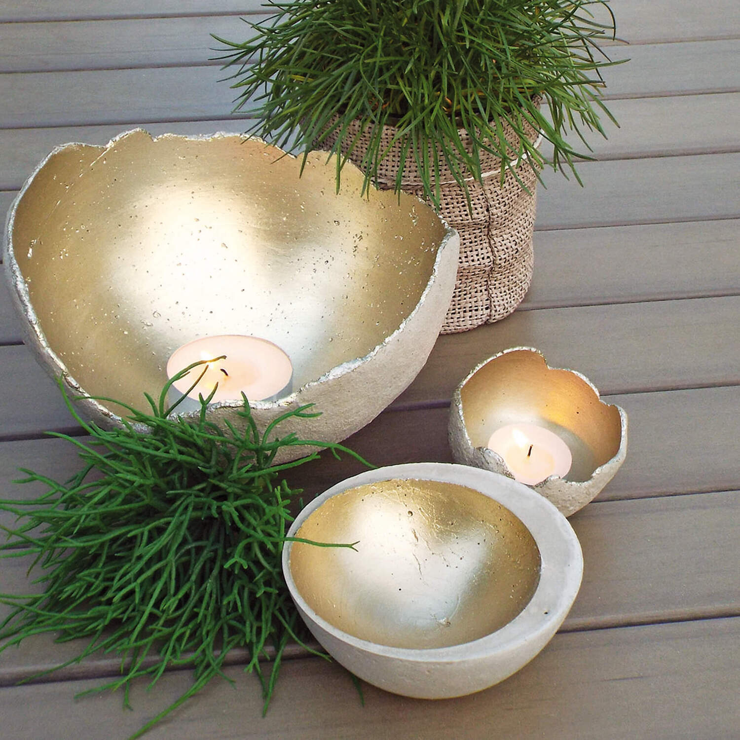 Bastelanleitung Deko-Schale aus Kreativbeton: Drei goldene Betonschalen mit Kerzen, umgeben von grünen Pflanzen auf einem Holztisch.