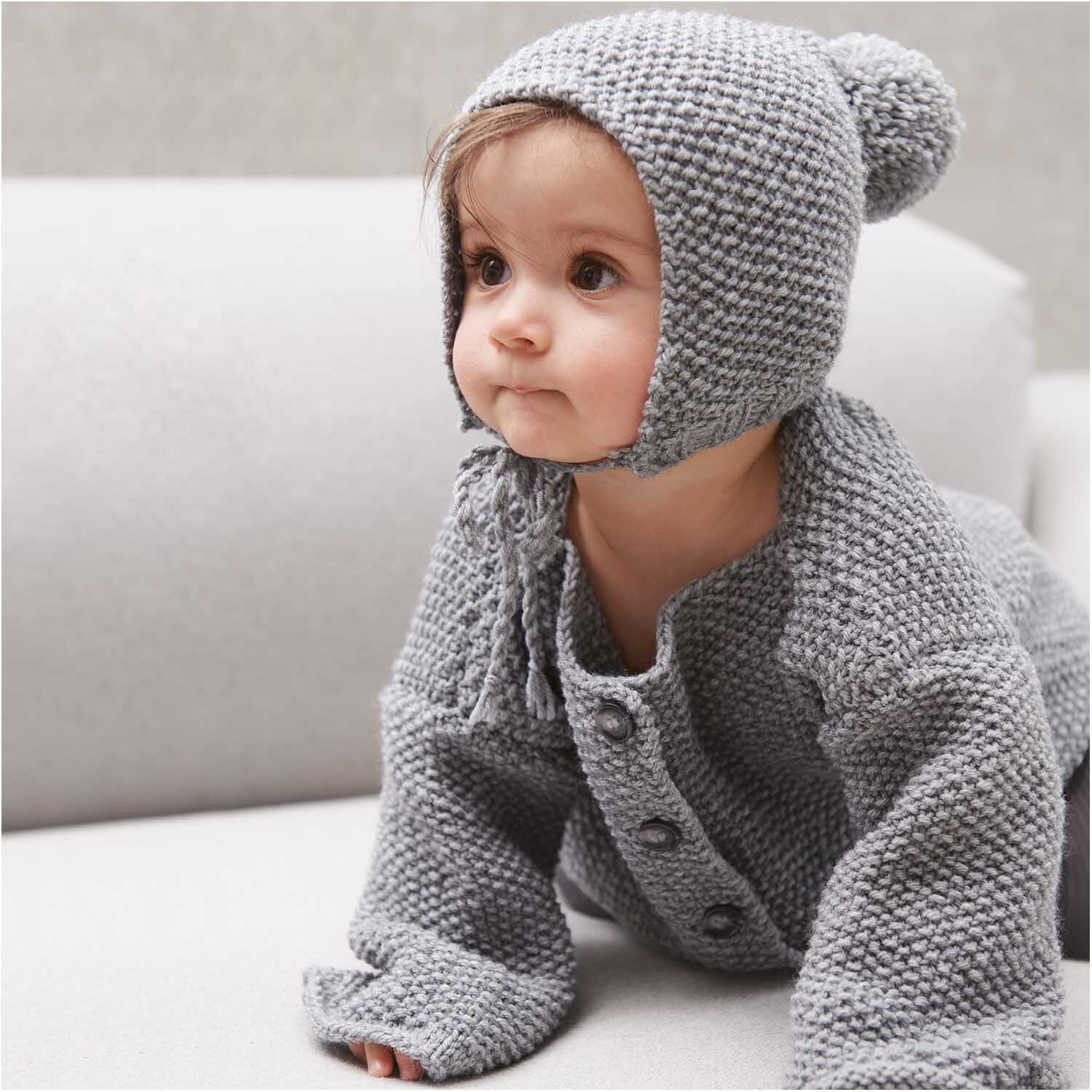 Rico Baby Classic dk: Ein Baby in gestrickter Kleidung aus silbernem Garn, auf einem Sofa sitzend.