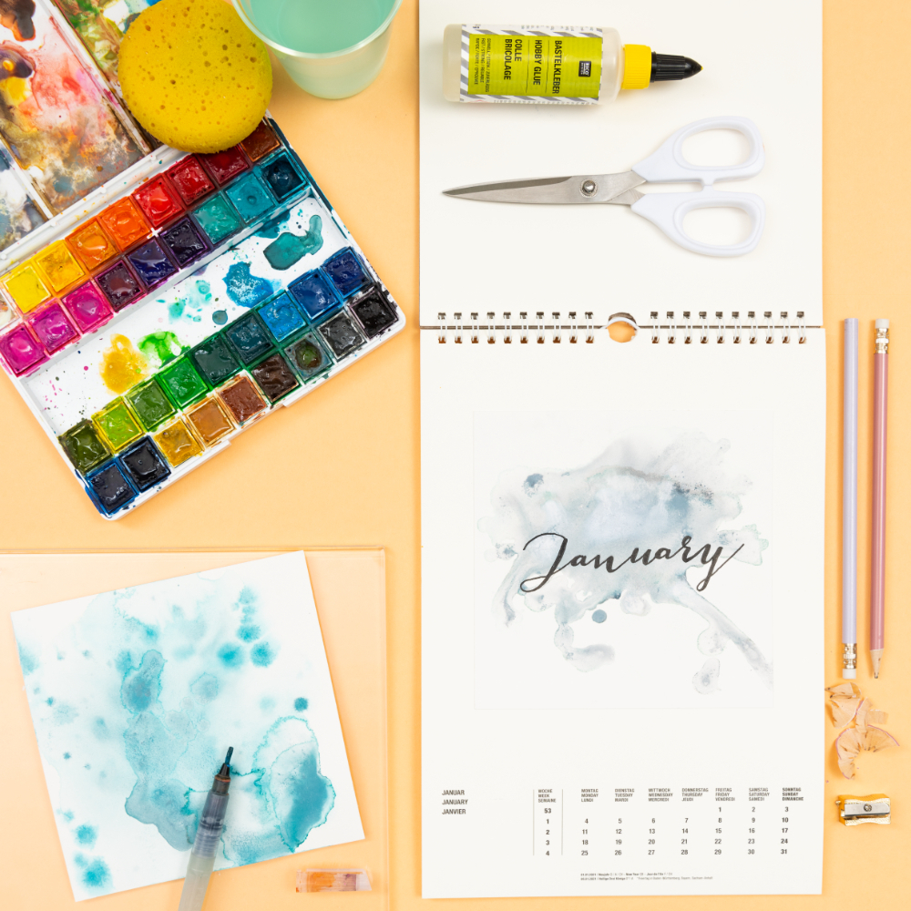 Anleitung Aquarell-Kalendergestaltung: Aquarellfarben, Pinsel, Schere und Kalenderblatt auf einem Tisch arrangiert.