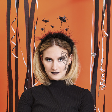 Frau mit Spinnennetz und schwarzen Augen geschminkt, dazu trägt sie einen Haarreif mit schwarzen Spinnen. Fotosetting orange Wand mit Luftschlangen.