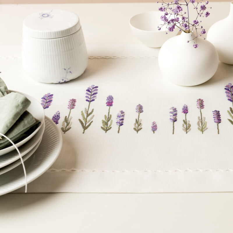 Stickpackung Läufer Lavendel zeigt zarte Lavendelstickerei auf Tischläufer, umgeben von weißem Geschirr. Ideal für elegante Tischdekoration.