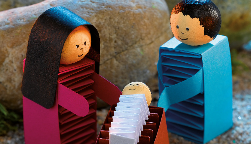 Bastelanleitung Weihnachtskrippe: Handgefertigte Figuren aus Papier in Blau und Rot vor einem Stein.